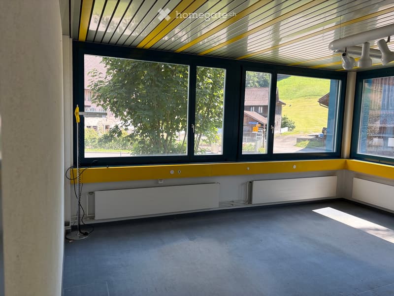 Empty room with large windows, yellow and white walls, grey carpeted floor, radiators, and a yellow ceiling with striped pattern.