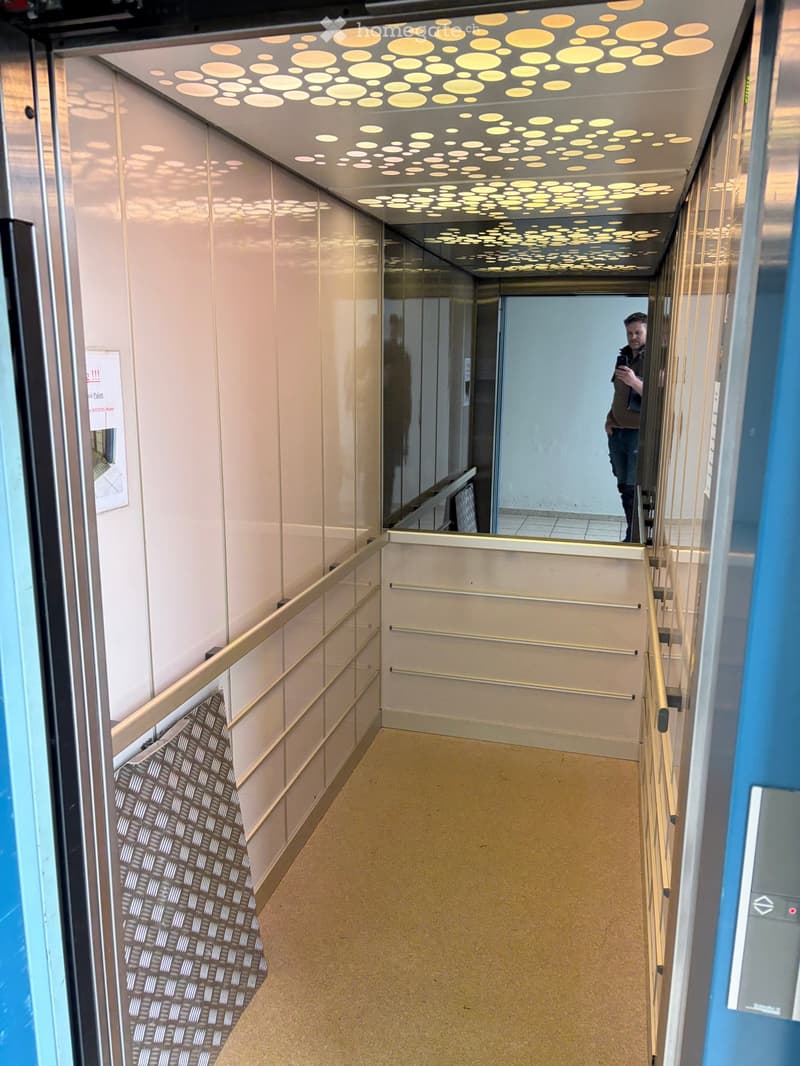 Modern hallway with light-colored walls, patterned carpet, and a decorative ceiling with circular light fixtures. Features a handrail and a glass wall on one side.