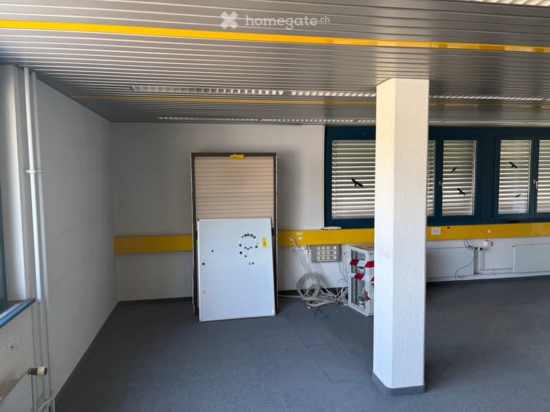 Empty room with grey concrete floor, white walls, and a central pillar. Features include a whiteboard, electrical panel, and a closed garage door.