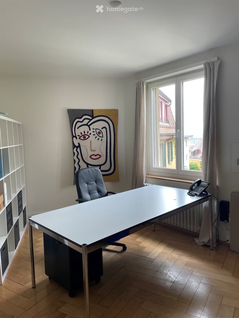 Office space with a large desk, office chair, telephone, shelf, and a large abstract painting on the wall. The room has wooden flooring and large windows with curtains.