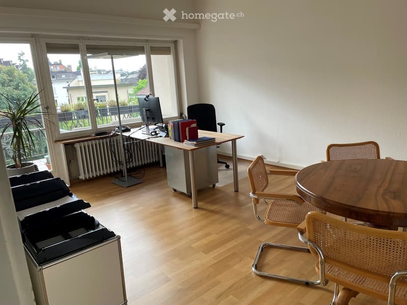 Office space with wooden flooring, large windows, a desk with a computer, a round table with chairs, and a potted plant near the window.