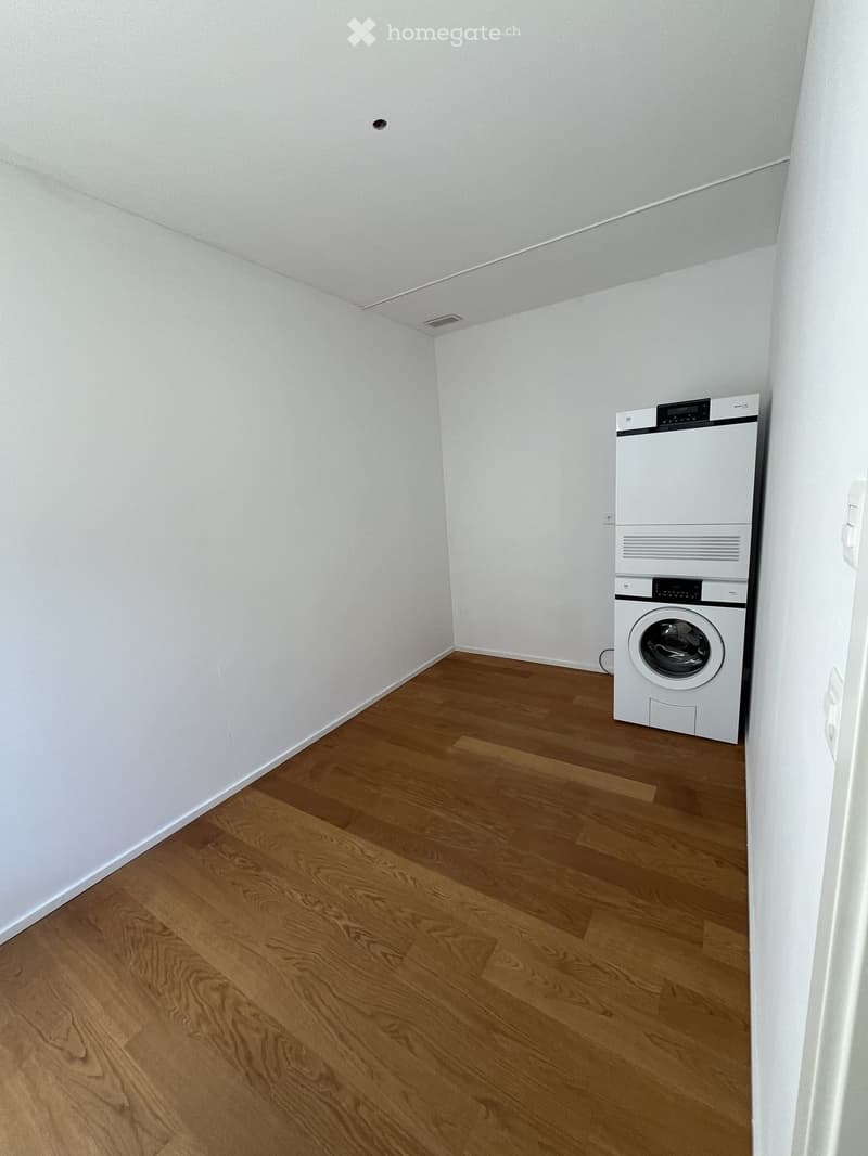 Small room with wooden flooring and white walls, featuring a stacked washing machine and dryer unit.