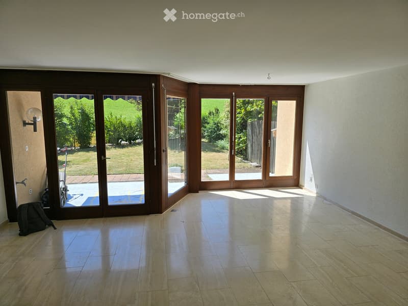 Empty room with tiled floor, large sliding glass doors leading to a garden, and a wall-mounted light fixture.