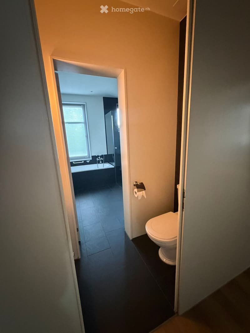 Bathroom with a toilet, bathtub, and a window with blinds. The floor is dark tiled, and the walls are light-colored.