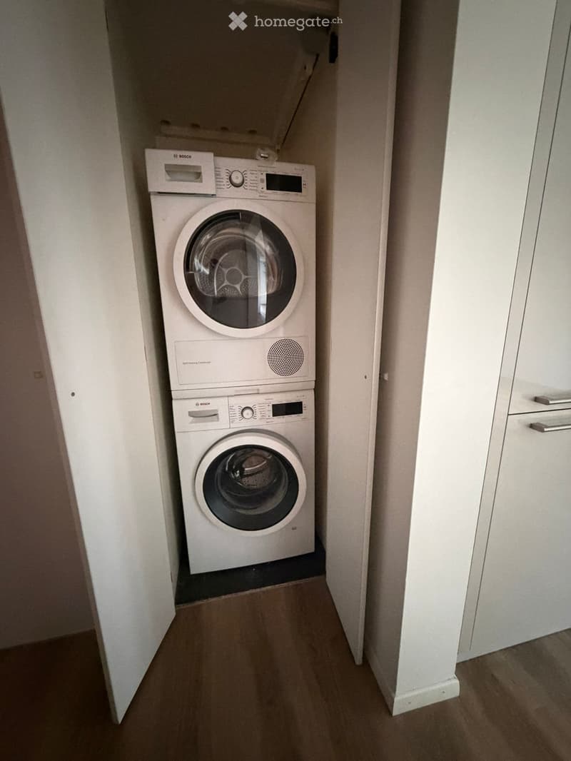 A small, well-lit room with a washing machine and a dishwasher stacked vertically, white walls, and wooden flooring.