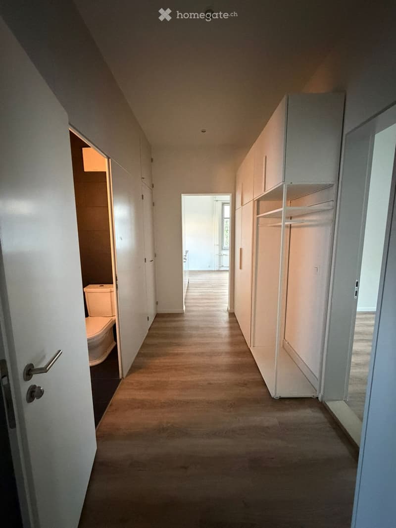 A narrow hallway with wooden flooring, white walls, and built-in storage units on one side. A partially open door on the left reveals a toilet, and another door at the end leads to another room.