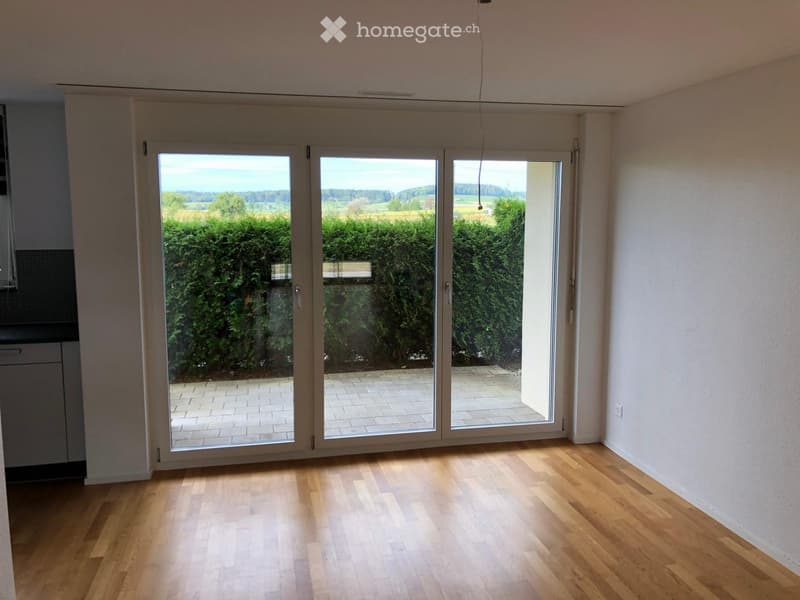 Empty room with wooden flooring and large sliding glass doors leading to a patio with a view of greenery and fields.