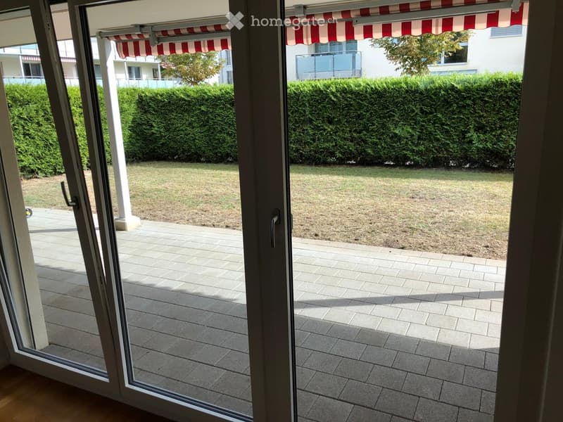 View from inside through large sliding glass doors onto a paved patio area with a small grassy lawn and trimmed hedges in the background.