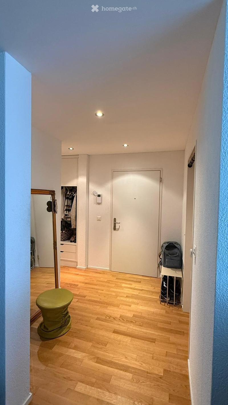 A narrow hallway with wooden flooring, a green stool, a mirror, a wardrobe, and a piece of luggage near the door.