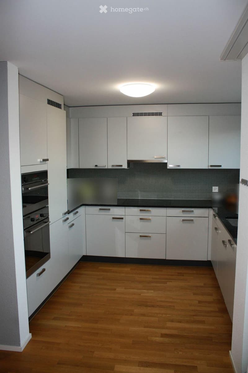 Modern kitchen with white cabinets, wooden flooring, and an integrated oven. The backsplash is dark grey tiles, and the countertops are black.