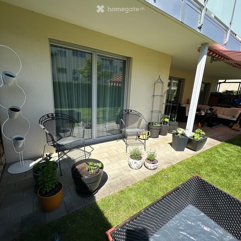 A well-maintained garden area with a tiled patio, potted plants, and outdoor furniture including chairs and a table.