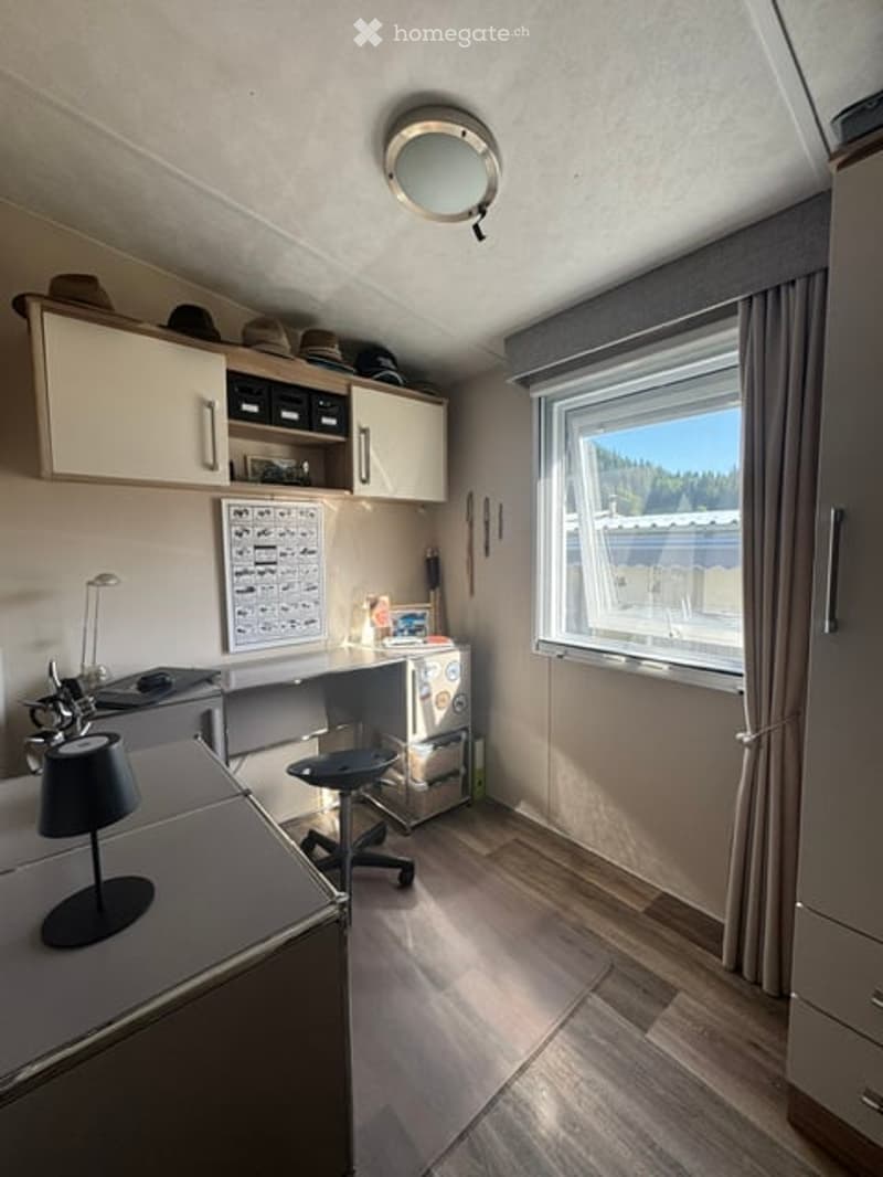 A modern, well-lit room with a desk, chair, lamp, and cabinet. The room features wooden flooring, a large window with blinds, and a round ceiling light fixture.