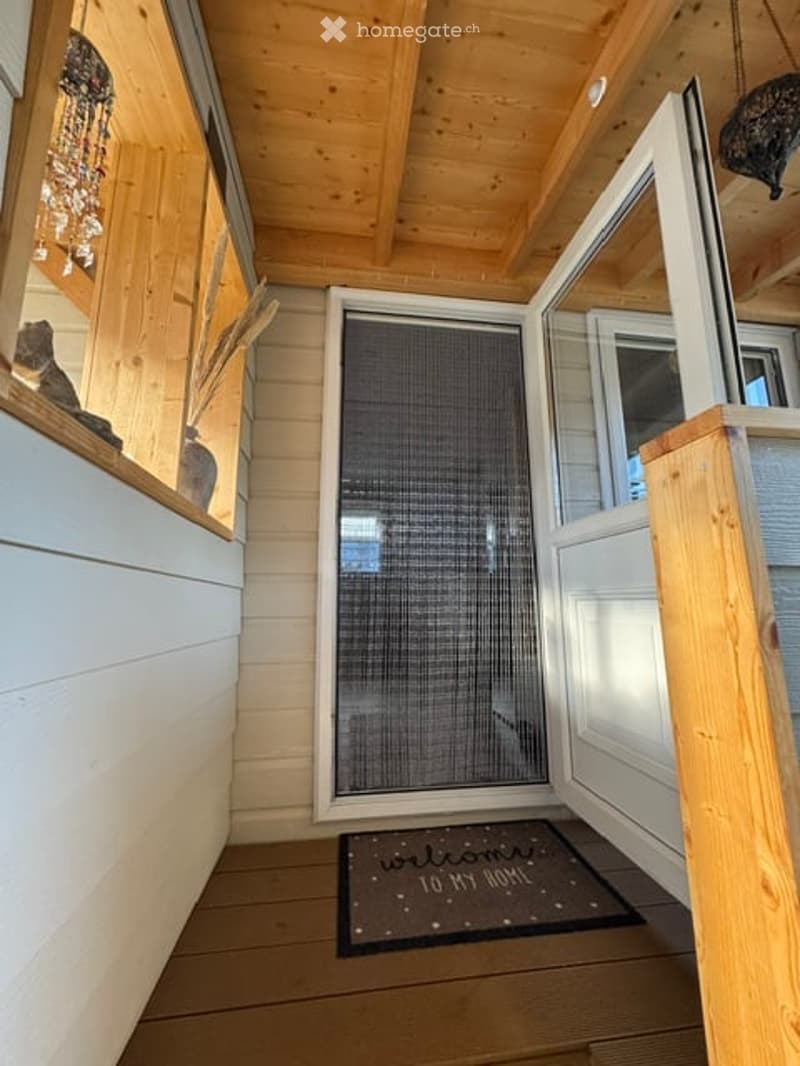A wooden-framed hallway with a glass door leading outside, a welcome mat, and decorative items on a shelf.