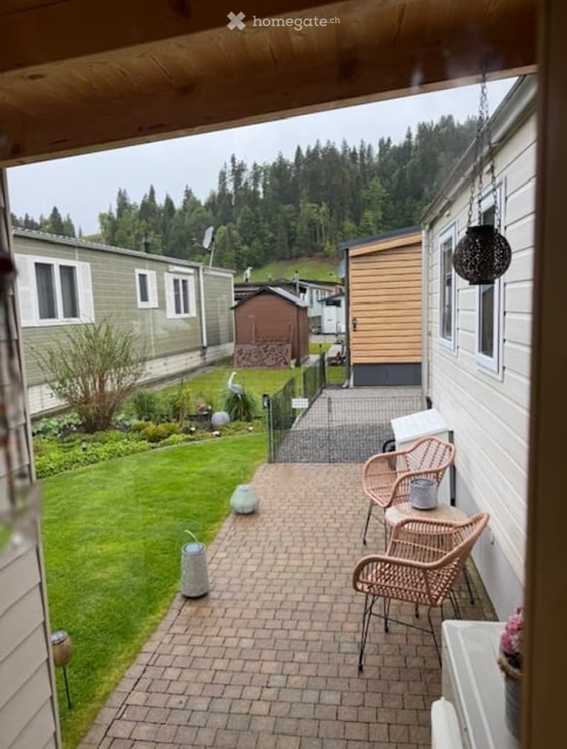 A well-maintained garden area with a brick-paved patio, outdoor furniture including wicker chairs and a small table, surrounded by green grass and trees.