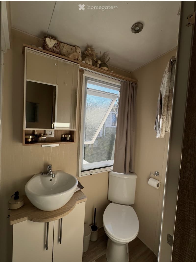 Compact bathroom with a modern sink, toilet, mirror cabinet, and window with a curtain. Includes a towel, toilet paper holder, and decorative items on the cabinet.
