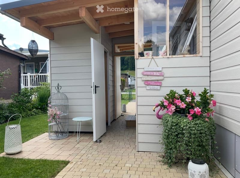 A cozy outdoor area with a brick pathway leading to a house entrance, featuring a small table, a birdcage with flowers, potted plants, and decorative signs on the wall.