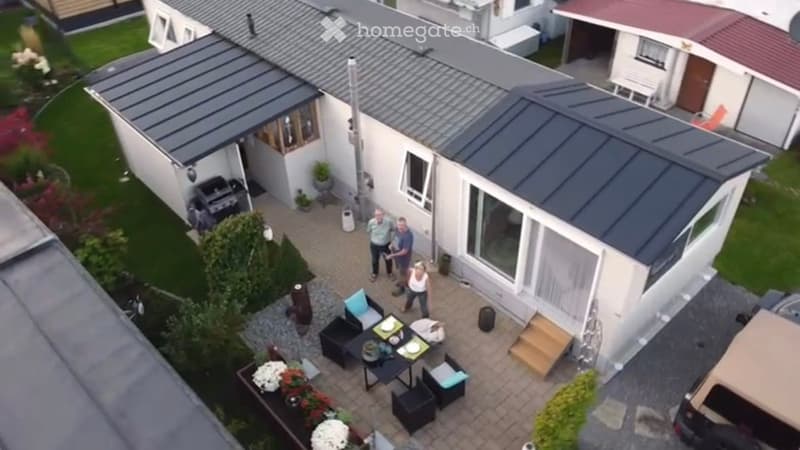Aerial view of a modern house with a patio area, outdoor furniture, and a small garden. The patio includes a dining table, chairs, and a grill, surrounded by greenery and neighboring houses.