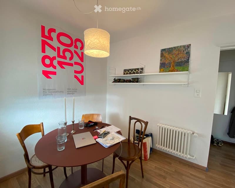 A small, well-lit room with a round wooden table surrounded by four wooden chairs. The table holds a laptop, a vase, and some papers. A white radiator is mounted on the wall, and a painting hangs above a shelf with small decorative items. A banner with te