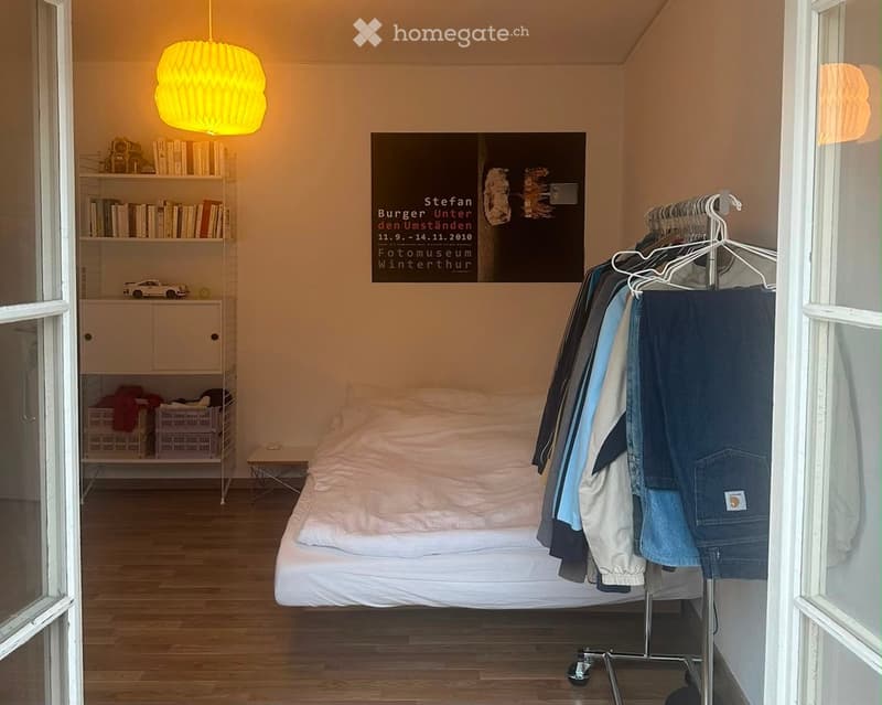 A small, modern bedroom with a single bed, a shelf with books and a toy car, a clothing rack with jackets, and a large picture frame on the wall.