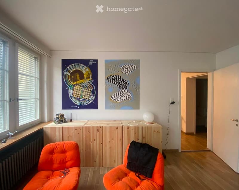 Bright living room with wooden flooring, two orange bean bag chairs, a small table with a lamp, and two posters on the wall. A doorway leads to another room.