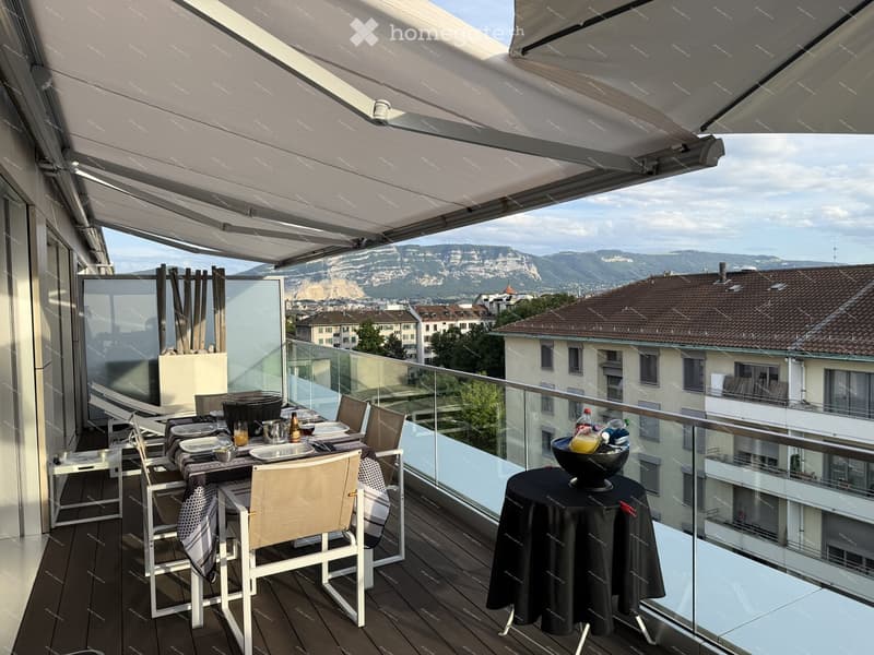 Balcony with a dining setup, including a table, chairs, and a bowl on a side table, overlooking a cityscape with mountains in the background.