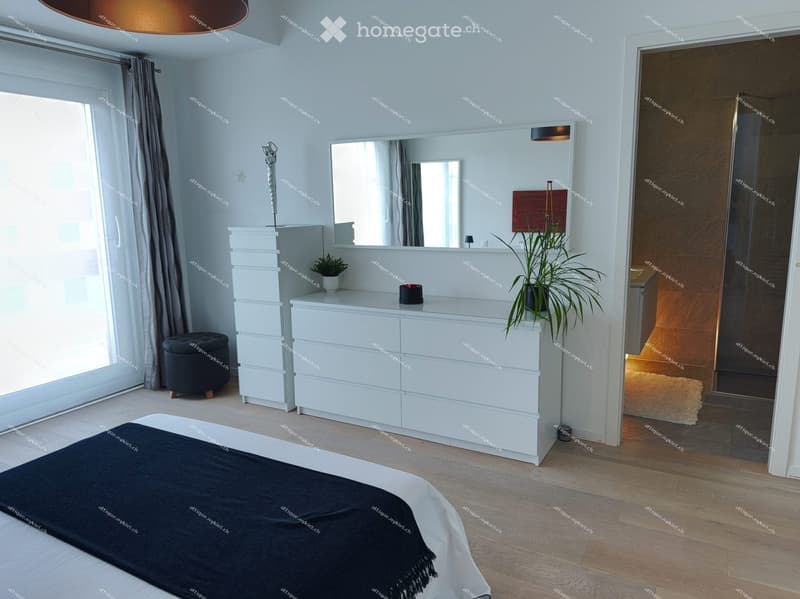 Modern bedroom with a large bed, white dresser, black stool, mirror, and decorative plants.