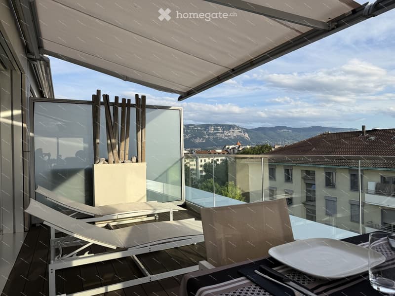 Modern balcony with a retractable awning, glass railing, lounge chairs, dining table, and scenic mountain view.