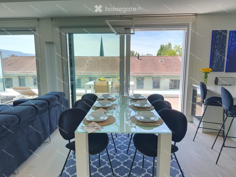 Modern dining and living area with a glass dining table, blue sofa, and large windows offering a scenic view of the city.