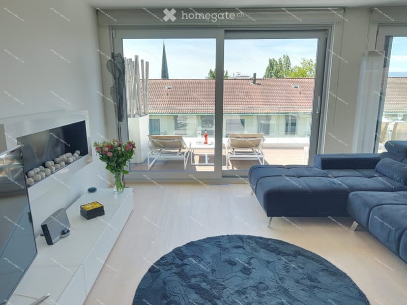 Modern living room with a dark blue sectional sofa, a white coffee table, a fireplace, a large window offering a view of the city, and a small outdoor seating area visible through the glass doors.