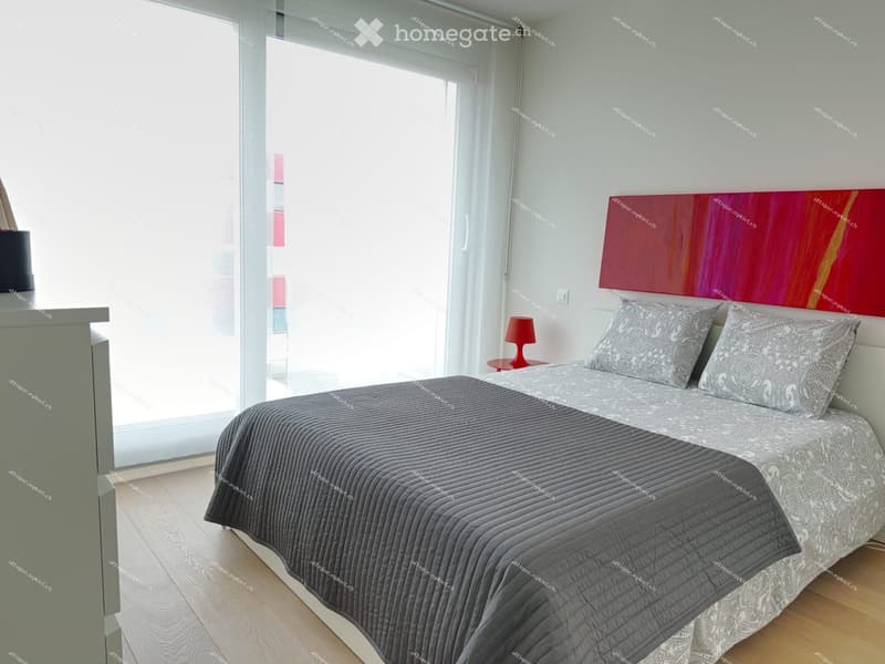 Modern bedroom with a double bed, grey bedding, nightstand with a red table lamp, and a colorful abstract painting on the wall.