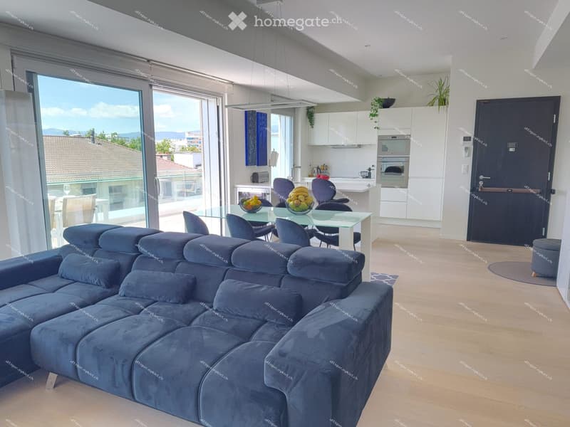 Modern living and dining area with a large sectional sofa, coffee table, dining table with chairs, and an open kitchen with white cabinets, an oven, and a refrigerator. The space features large windows with a view of the city and mountains.
