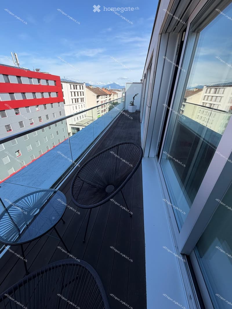 Modern balcony with glass railing, wooden deck, and cityscape view. Includes a round glass table and two black wire chairs.