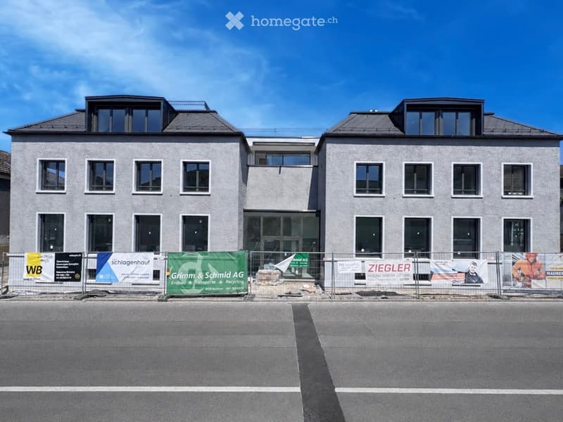 Building. Two-story building with modern facade, multiple windows, and a flat roof with small dormers. The building is surrounded by a temporary fence with various advertisements.