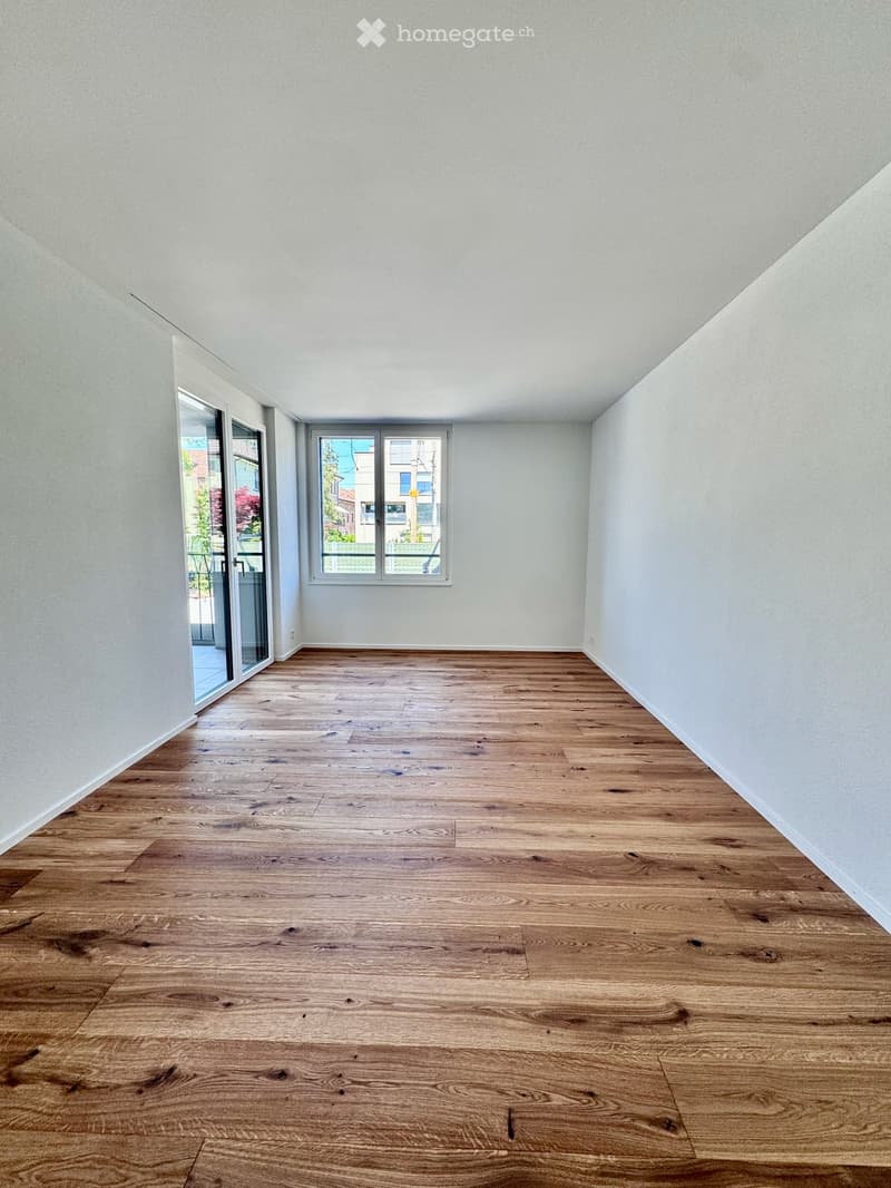 Empty room with wooden flooring and white walls, featuring a large window and a sliding glass door leading outside.