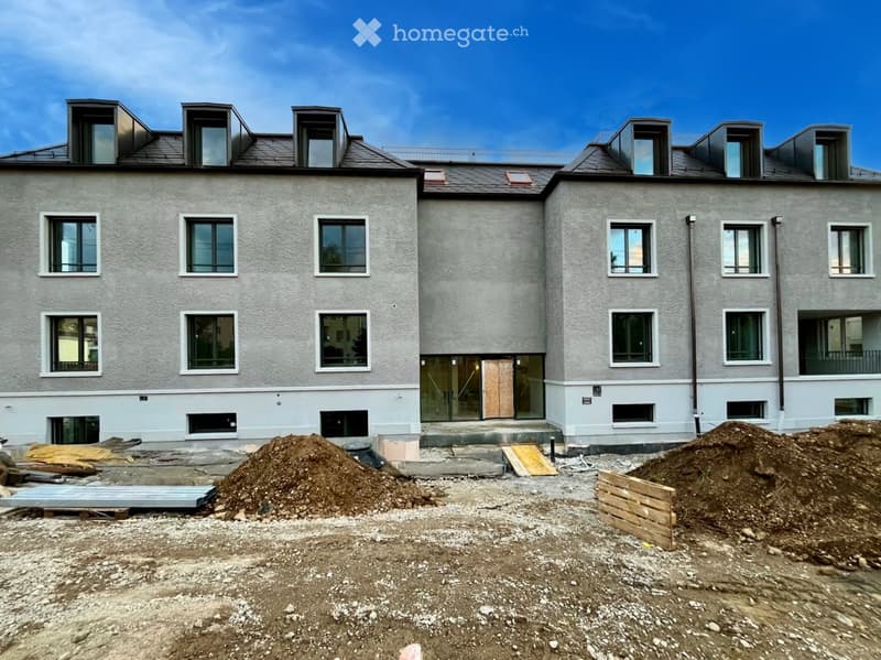 Building. Two-story residential building with grey facade, multiple windows, and a modern design. The ground level shows construction materials and dirt piles.
