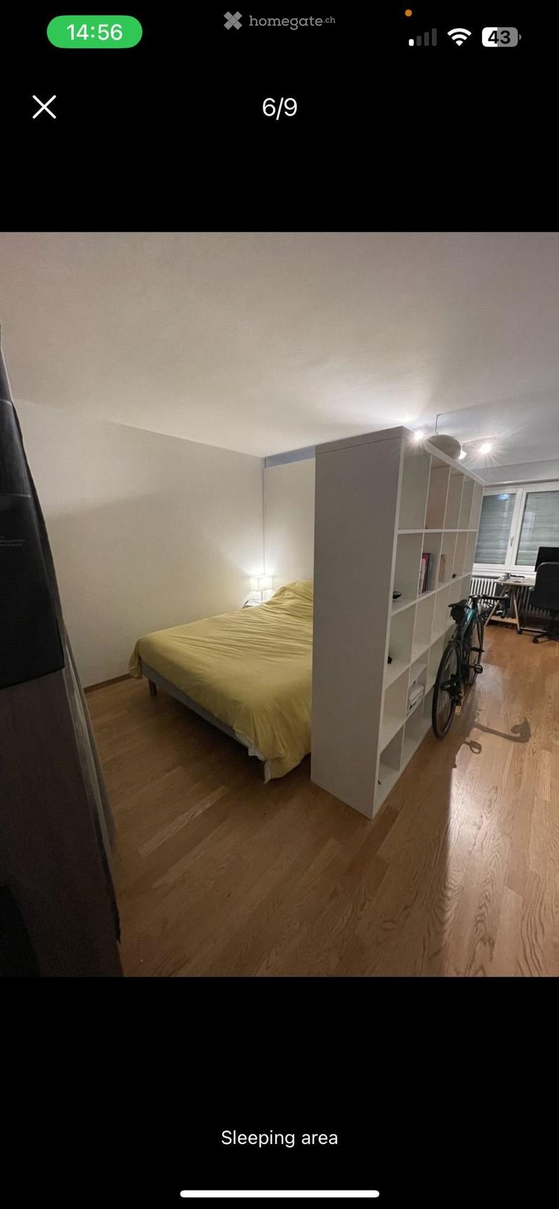 A modern bedroom with a wooden floor, a bed with yellow bedding, a nightstand with a lamp, a bookshelf, and a bicycle leaning against it.