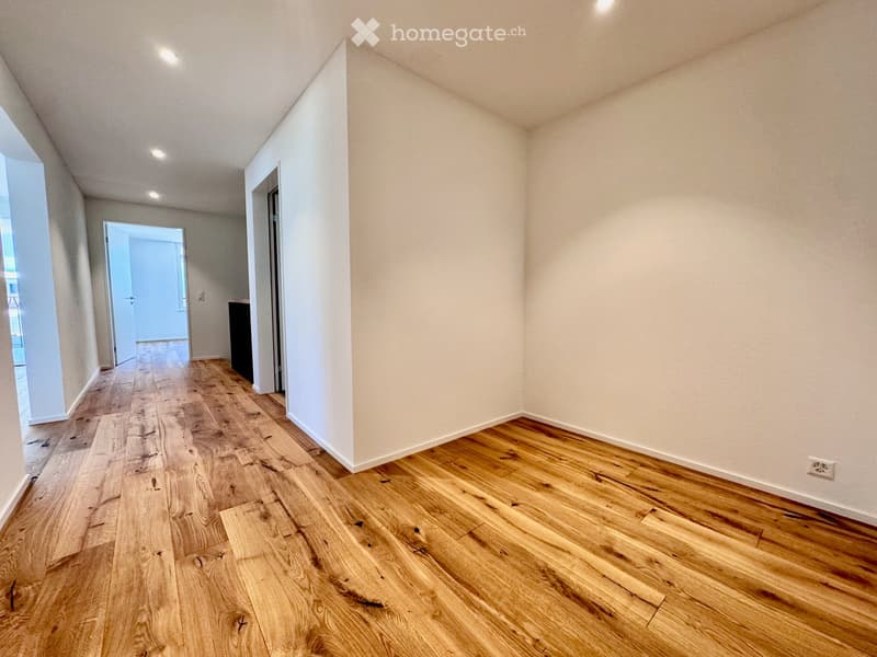 Empty hallway with wooden flooring and white walls, featuring recessed lighting and an open doorway leading to another room.