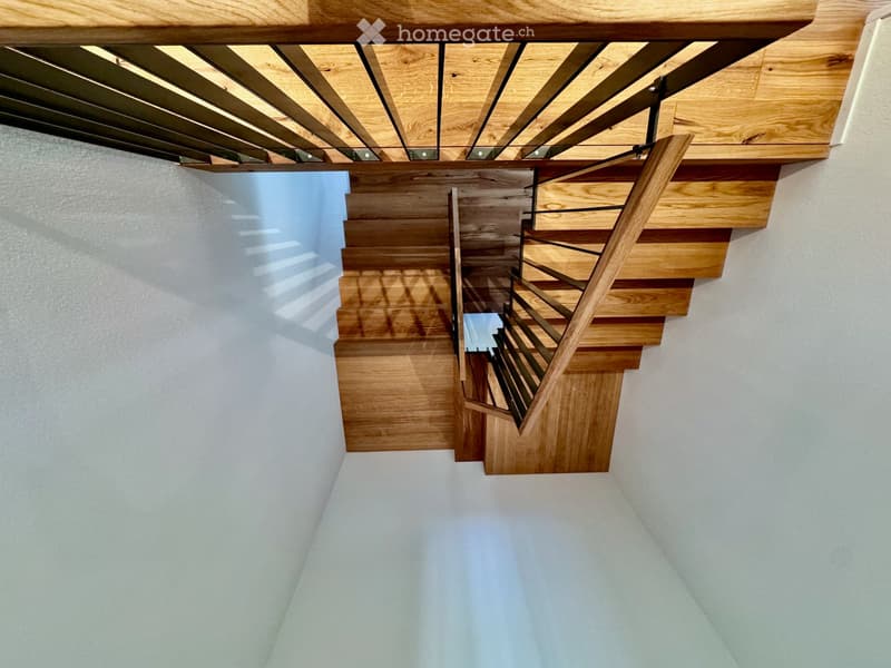 Modern wooden staircase with open wooden railings and a glass ceiling, leading upwards in a bright, minimalist hallway.