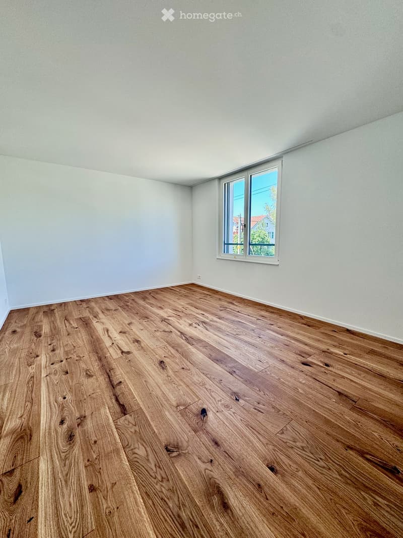 Empty room with light-colored walls and a large window, featuring a polished wooden floor.