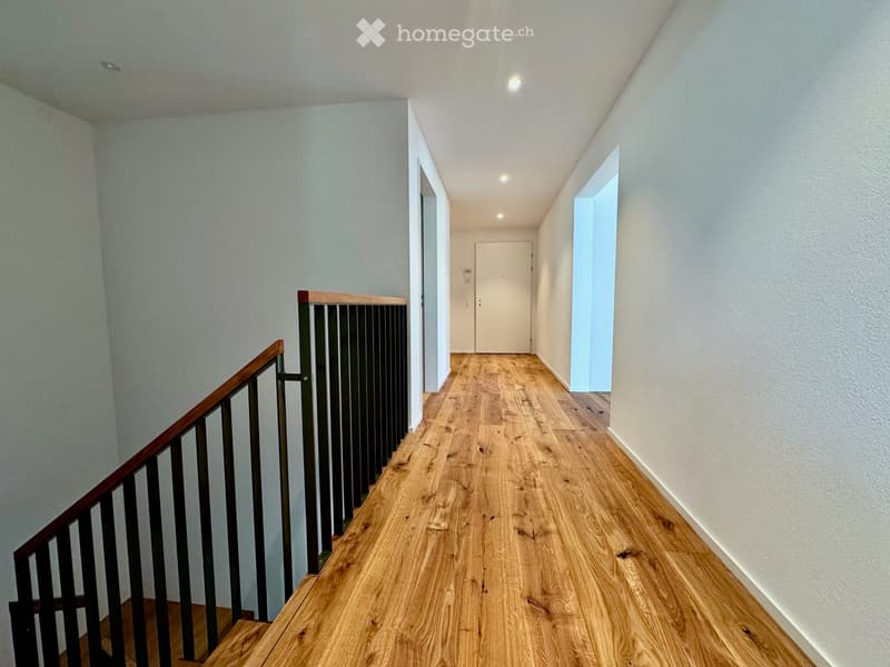 A modern hallway with wooden flooring, white walls, and a staircase with a wooden handrail and metal balustrade. The hallway features recessed lighting and a closed door at the end.