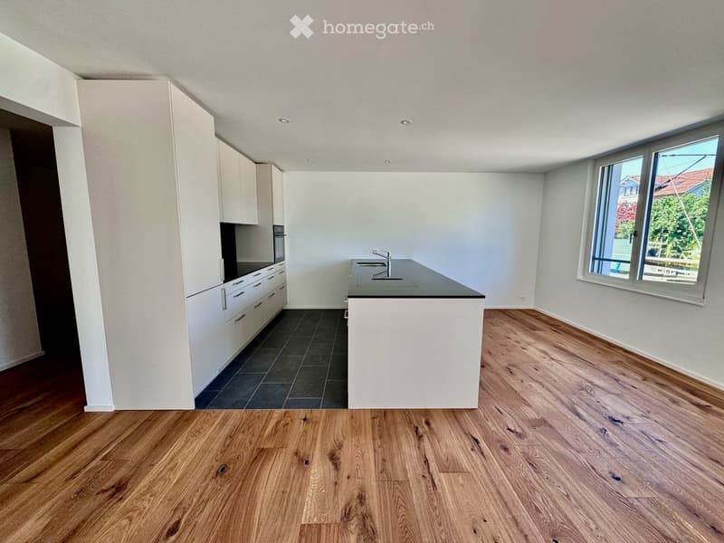 Modern kitchen with white cabinets, a central island, and a dark countertop. The floor is made of light wooden planks, and there is a large window on the right side.