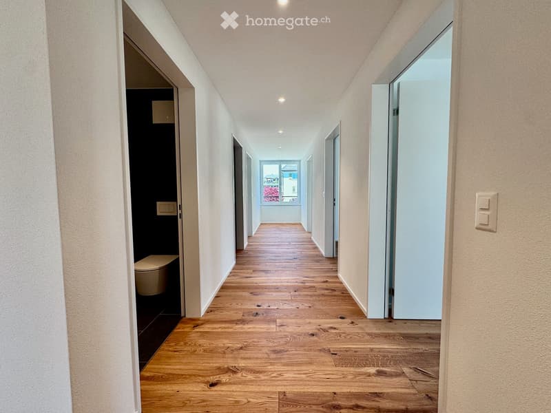 A long, narrow hallway with wooden flooring, white walls, and recessed lighting. Doors lead to various rooms, including a bathroom with a toilet visible on the left side.