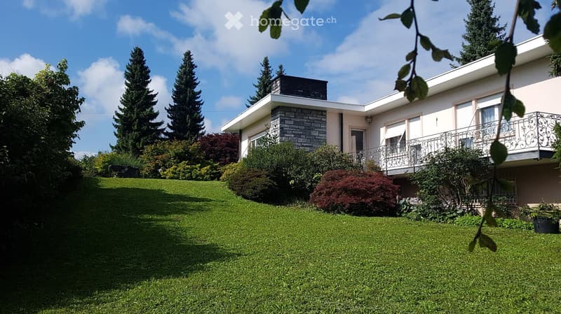 A two-story house with a modern design, featuring a raised ground floor, a balcony, and a well-maintained garden with green lawns and various trees and shrubs.