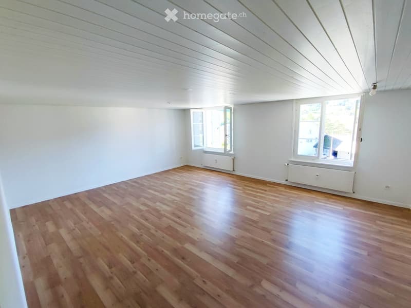 Empty room with wooden flooring, white walls, and large windows allowing natural light. The ceiling has a slanted design typical of an attic space.