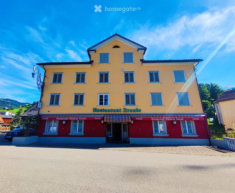 Two-story yellow building with white trim, red awning, and signage reading "Restaurant Pizzeria Traube".