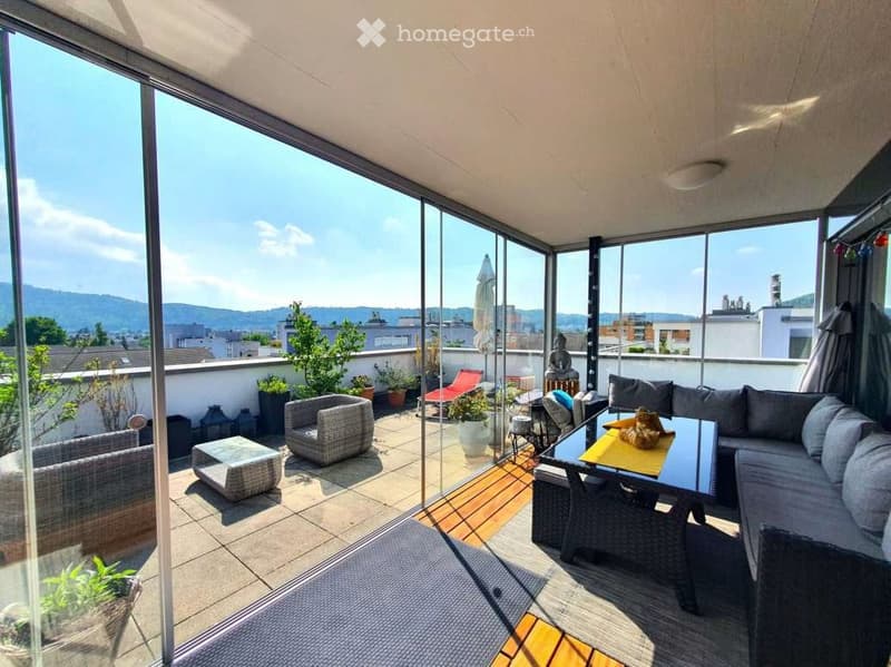 Modern balcony with large glass walls, offering a scenic city view. Features a grey sofa, a coffee table with a yellow runner, wicker chairs, a red lounge chair, potted plants, and a decorative statue.