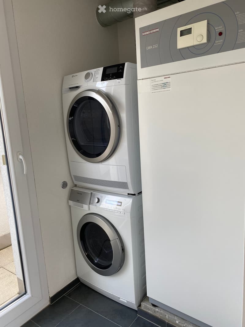 A small utility space with a washing machine, dryer, and water heater. The appliances are white and modern, with control panels visible.