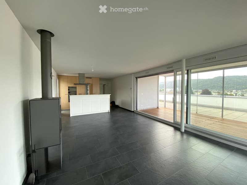 Open-plan living space with dark tiled floor, white kitchen island, wooden deck, and large sliding glass doors leading to a balcony with scenic mountain view.
