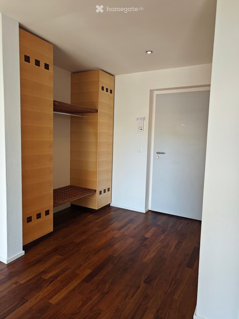 Empty room with wooden flooring, white walls, and a built-in wooden closet with shelves. A closed door is visible on the right side.