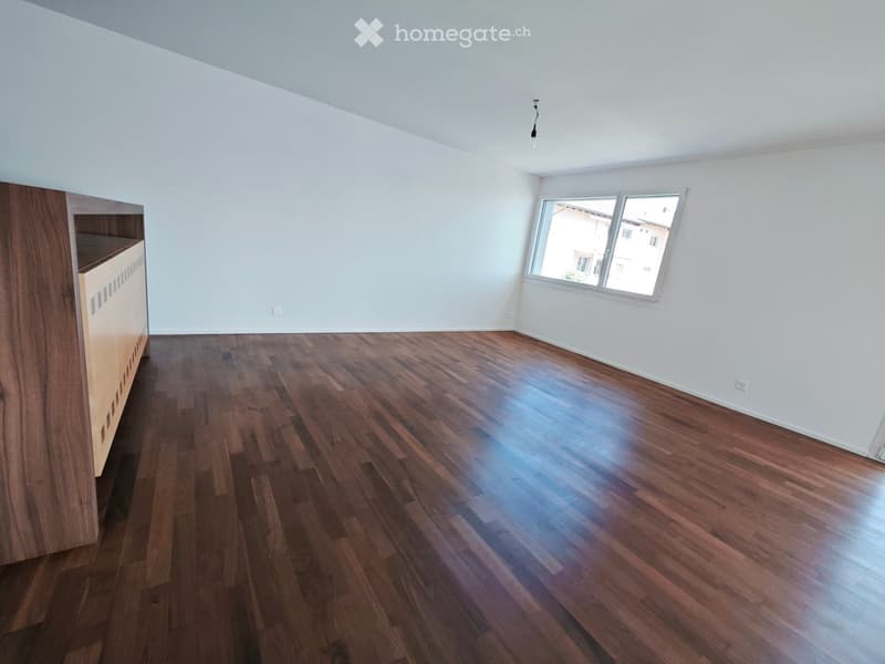 Empty room with wooden flooring, white walls, and a window letting in natural light. A small wooden cabinet is visible on the left side.